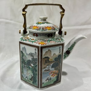 Könnte beinhalten: Eine weiße Porzellan-Teekanne mit goldenem Griff und Ausguss. Die Teekanne hat handgemalte Landschaftsszenen auf jeder Seite und einen dekorativen Deckel. Der Griff ist aus goldfarbenem Metall. Eine einzigartige Dekoration im asiatischen Stil.