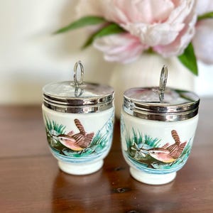 May include: Two white ceramic egg coddlers with silver-toned metal lids featuring a ring handle. Each coddler is decorated with a colorful illustration of a small brown bird perched among green reeds and blue water accents on a white background.