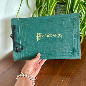 May include: A rectangular, dark green photo album with a textured cover featuring an embossed border design and the word "Photographs" printed in gold lettering. The album is bound on the left side with a black cord tied through two metal eyelets.