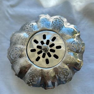 May include: A silver-coloured, ornate drain cover with a floral and scrollwork design. The centre features a circular, perforated plate with a central knob. The rim has a scalloped edge, and the overall aesthetic is vintage.
