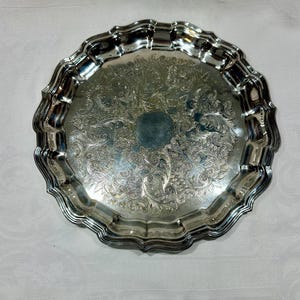 May include: A silver-coloured, round serving tray with a scalloped edge. The tray features an ornate, engraved floral pattern across its surface. The metal reflects light, highlighting the intricate details of the design. The tray is likely made of silver or a silver-plated material.