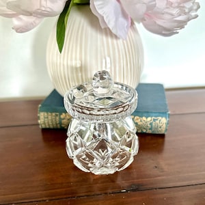 Könnte beinhalten: Ein klares, facettiertes Glasgefäß mit passendem Deckel. Das Gefäß hat einen runden Boden und ein Rautenmuster. Der Deckel hat einen kleinen, runden Knopf. Das Gefäß steht auf einer Holzoberfläche, mit einer weißen Vase und rosa Blumen im Hintergrund.