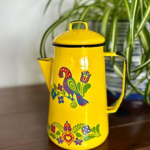 Puede incluir: Una cafetera de esmalte amarillo brillante con asa y tapa negras. La cafetera está decorada con un diseño floral y de pájaro colorido. La cafetera mide aproximadamente 23 cm de altura.