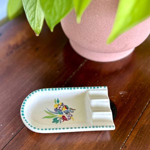 Può includere: Un posacenere vintage in ceramica bianca con un motivo floreale e di uccelli. Il posacenere ha una forma rettangolare con bordi arrotondati, tre posa-sigarette e un bordo a pois turchese. Il motivo floreale presenta fiori gialli, blu e rossi.