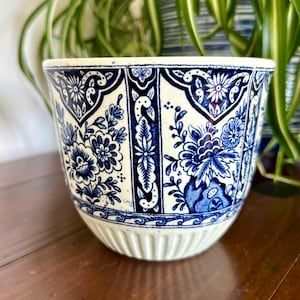 May include: A ceramic planter featuring a white base with intricate blue floral patterns and geometric borders. The upper section displays detailed botanical illustrations, while the lower portion has a subtle vertical ribbed texture.