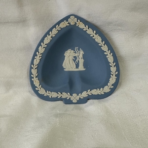 May include: A heart-shaped, blue ceramic dish with a white, raised relief design. The dish features a white floral and leaf border. The centre of the dish has a white relief of classical figures. A decorative piece for display.