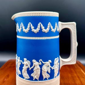 May include: A blue ceramic pitcher with white relief detailing. The pitcher features a garland design around the top and bottom, and a frieze of dancing figures. The handle and rim are white.