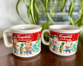 May include: Two vintage Campbell's soup mugs. Each mug features a red band with the Campbell's logo and the phrase "M'm! M'm! Good!". The mugs have a white background with cartoon illustrations of children in a garden.