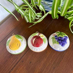May include: Three ceramic fruit-themed wall hangings. The set includes a yellow pear, a red apple, and a cluster of purple grapes. Each piece has a white background with green leaves and a gold hanging loop. The edges are trimmed with color.