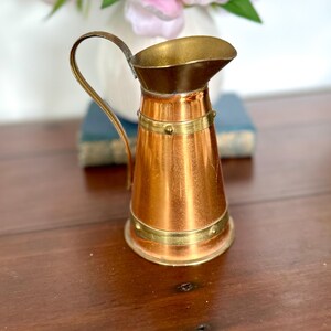 May include: A small copper jug with a brass handle and rim. The jug has a conical shape with brass bands around the top and bottom. The handle curves upwards. The jug is on a wooden surface.