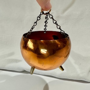 May include: A small, round copper cauldron with three short legs and a chain for hanging. The cauldron has a polished, reflective surface and a dark interior. The chain is made of dark metal links and is attached to a ring at the top.