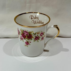 Può includere: Tazza in ceramica bianca con bordo e dettagli del manico dorati. La tazza è decorata con un motivo floreale di rose rosa e foglie verdi. Le parole "Ruby Wedding" sono scritte all'interno del bordo.