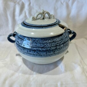 May include: A ceramic soup tureen with a lid. The tureen is round with a blue speckled pattern on the bottom half and white on the top half. It has two handles and a lid with a decorative handle.