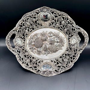 Puede incluir: Un bol de servir de plata con un diseño ornamentado que presenta un motivo floral y un diseño central de frutas. El bol tiene dos asas y está hecho de metal plateado.