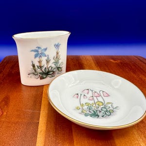 Pode incluir: Dois pratos de porcelana branca com acabamento dourado. O prato maior tem um desenho floral com flores cor-de-rosa e folhas verdes. O prato menor tem um desenho floral com flores azuis e folhas verdes.