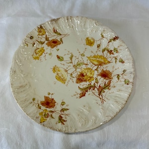 May include: An antique cream-coloured plate with a scalloped edge. The plate features a floral design in shades of yellow, orange, and brown. The flowers are arranged in a delicate pattern across the surface, creating a vintage aesthetic.