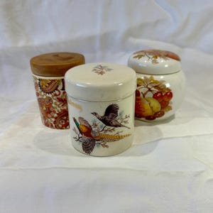 May include: Three decorative ceramic jars with lids. One jar has a wooden lid and a red and orange paisley pattern. Another jar features a pheasant design, and the third jar has a fruit design. The jars are on a white surface.