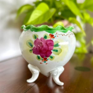 May include: A white ceramic planter with a frilled green rim and three small feet. The planter features a large pink rose and green leaves. The background shows blurred green foliage.