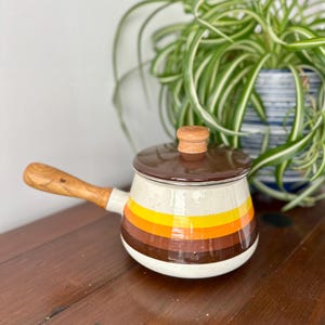 May include: A small enamel saucepan with a speckled cream base featuring horizontal stripes in yellow, orange, and brown. It includes a brown lid with a light wood knob and a matching light wood handle extending from the side of the pot.