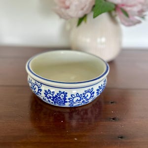 May include: A small, round ceramic bowl with a cream-coloured interior and a white exterior featuring a repeating blue floral pattern. The rim of the bowl is accented with a thin blue line, and the base is slightly tapered with a smooth finish.