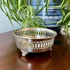 Puede incluir: Un cuenco redondo de metal plateado con un intrincado diseño de celosía calada a lo largo del borde superior. El cuenco se apoya sobre tres pies pequeños y ornamentados y tiene una superficie interior lisa y reflectante.