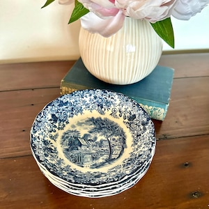 May include: A stack of small, blue and white decorative plates with a landscape design. The plates have a floral border and a central scene of a house, trees, and water. A white vase with pink flowers is in the background.