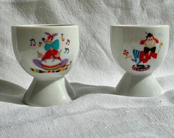 May include: Two white ceramic egg cups with colorful cartoon illustrations. One features a character playing a drum, the other a clown. Both have a pedestal base and are likely vintage collectibles.