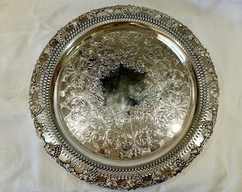 May include: A silver-colored, round serving tray with intricate floral and scrollwork patterns. The tray has a detailed, pierced border with grape cluster designs. The center of the tray reflects light, highlighting the ornate details.