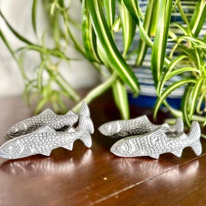 Puede incluir: Cuatro objetos decorativos plateados con forma de pez están dispuestos sobre una superficie de madera. Los peces tienen escamas y aletas detalladas. Plantas verdes y blancas en el fondo añaden un elemento natural.