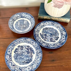 Puede incluir: Tres platos decorativos azules y blancos con diseños florales y paisajísticos. Los platos presentan un detallado patrón de transferencia azul sobre fondo blanco. Los platos están dispuestos sobre una superficie de madera.