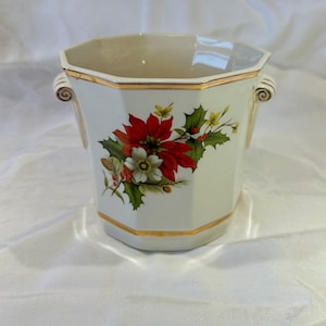 Könnte beinhalten: Eine weiße, achteckige Keramikvase mit goldenen Verzierungen und Griffen. Die Vase hat ein Blumendesign mit roten Weihnachtssternen, weißen Blüten, grünen Blättern und Beeren. Die Vase ist ein dekorativer Gegenstand.