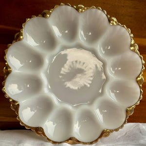 May include: A white, scalloped egg plate with a gold-colored rim. The plate has 12 egg indentations and a central well. The gold rim adds a touch of elegance to the vintage-style serving dish.