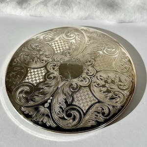 May include: A silver-toned, round compact mirror with intricate floral and scrollwork engraving. The design includes a central, unadorned circle and patterned diamond shapes. The compact has a reflective, polished surface.