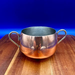 May include: A copper cup with two curved handles. The cup has a shiny, reflective surface and a silver interior. The cup is on a wooden surface with a blue background.