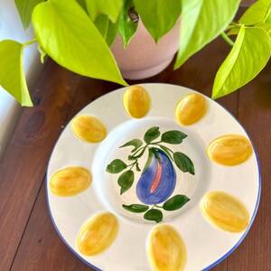 May include: A white ceramic serving plate with a blue rim and a central design of a blue eggplant with green leaves. The plate has eight yellow egg-shaped indentations around the edge.