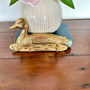 May include: A gold-colored duck-shaped wall hook. The duck is detailed with textured feathers and a long neck. The hook is resting on a blue book, with a white vase and pink flowers in the background. The hook is approximately 6 inches long.