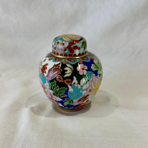 Op de afbeelding: Een kleine, decoratieve pot met deksel, met een kleurrijk bloemmotief. De pot heeft een gouden rand en basis, met een mix van blauwe, rode, witte en roze bloemen. Het deksel is ook versierd met bloemen en gouden accenten.