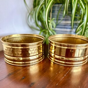 Könnte beinhalten: Zwei runde Pflanzgefäße aus poliertem Messing mit glänzendem Metallfinish. Das linke Gefäß weist eine texturierte, gehämmerte Oberfläche auf, während das rechte Gefäß eine glatte Oberfläche hat. Beide Behälter haben ein einfaches, offenes Design.