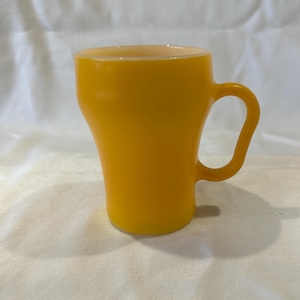 Puede incluir: Una taza amarilla con un asa curva. La taza tiene una forma ligeramente acampanada, más ancha en la parte superior que en la base. La taza está hecha de un material sólido y opaco y se encuentra sobre una superficie blanca.