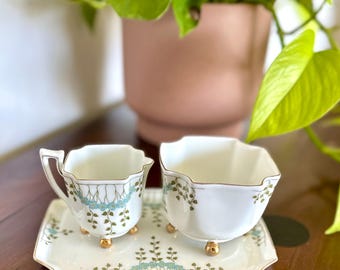 May include: A vintage white teacup and sugar bowl set with gold trim and delicate blue and green floral accents. The set includes a rectangular tray with matching design, all resting on small gold feet. The teacup has a handle.