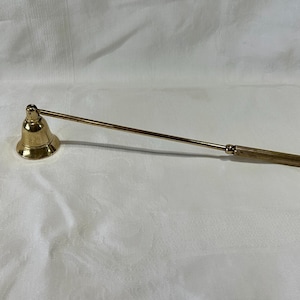 May include: A gold-colored candle snuffer with a bell-shaped head and a long, slender handle. The handle has a textured grip for easy handling. The snuffer is designed to extinguish candles safely.