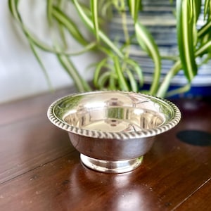 Op de afbeelding: Een kleine, glanzende zilveren kom met een decoratieve, touwachtige rand. De kom staat op een korte, ronde voet. De kom is leeg en weerspiegelt de omgeving. De kom is waarschijnlijk gemaakt van metaal en is geschikt voor het serveren van kleine porties.