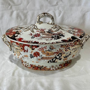 Può includere: Una zuppiera in ceramica bianca e oro con coperchio. Il piatto presenta un design floreale e paesaggistico rosso, nero e oro. Il coperchio ha una maniglia e una fessura per un cucchiaio. Il piatto ha due maniglie laterali.