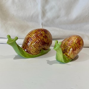 May include: Two ceramic snail ornaments. The snail shells are brown with yellow and red stripes. The snail bodies are green. The snails are on a white surface.
