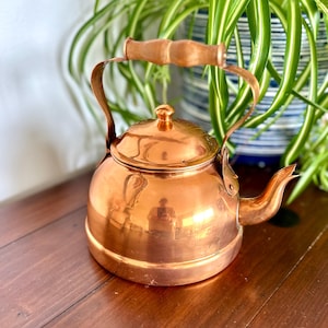 May include: A copper tea kettle with a wooden handle and lid knob. The kettle has a rounded body and a curved spout. The copper surface reflects the surrounding environment. The handle is attached to the kettle with copper brackets.