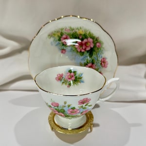 Peut inclure: Un service à thé vintage avec un motif floral. La tasse et la soucoupe en porcelaine blanche présentent des fleurs roses et des feuilles vertes avec une bordure dorée. La tasse repose sur un support doré.
