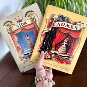 Könnte beinhalten: Zwei Vintage-Bücher mit den Titeln "Aida" und "Carmen". Das "Aida"-Buch hat einen hellbeigen Einband mit einer Bühnenillustration. Das "Carmen"-Buch hat einen gelben Einband mit einer roten Bühnenillustration. Beide Bücher stammen von The Metropolitan Opera Guild.