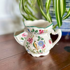 Puede incluir: Jarra de afeitar de cerámica color crema con diseños florales y de pájaros. La jarra tiene un asa y un pico, con un borde verde. El patrón floral incluye flores rosas, azules y amarillas. Un artículo decorativo.