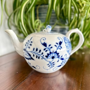 Może przedstawiać: Biały porcelanowy czajniczek z niebieskim wzorem cebulowym oraz misternymi motywami kwiatów i pnączy. Czajniczek posiada zakrzywioną wylewkę, zaokrąglony korpus, dopasowaną pokrywkę z uchwytem w kształcie gałki oraz solidną rączkę z boku.