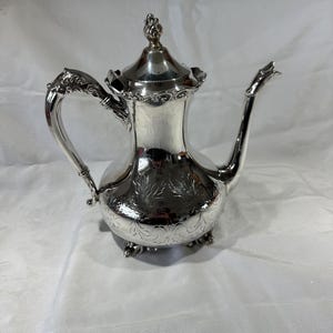May include: A silver-colored teapot with a decorative floral design. The teapot has a curved handle, a spout, and a lid with a finial. The teapot is resting on a white surface.